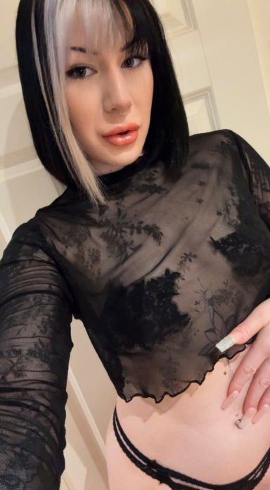 Kvinna i genomskinlig blus och underkläder, poserar mot vit bakgrund.